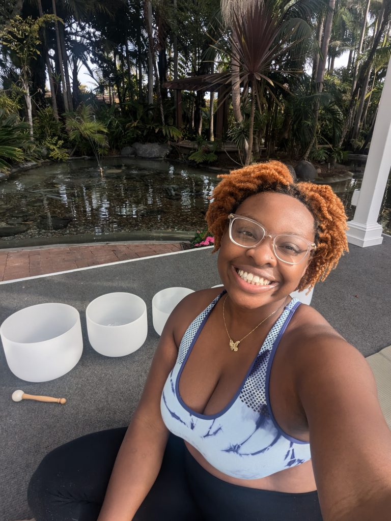 Pictured in NeNe smiling into the camera with white, crystal sound bowls behind her. In the distance is a body of water and palm trees. NeNe found comfort from the pain of death in the magick of sound bowls. 