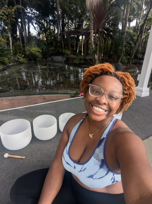 NeNe sits in front of a small pond lined with greenery. She smiles in a selfie sitting on the floor with her sound bowls.