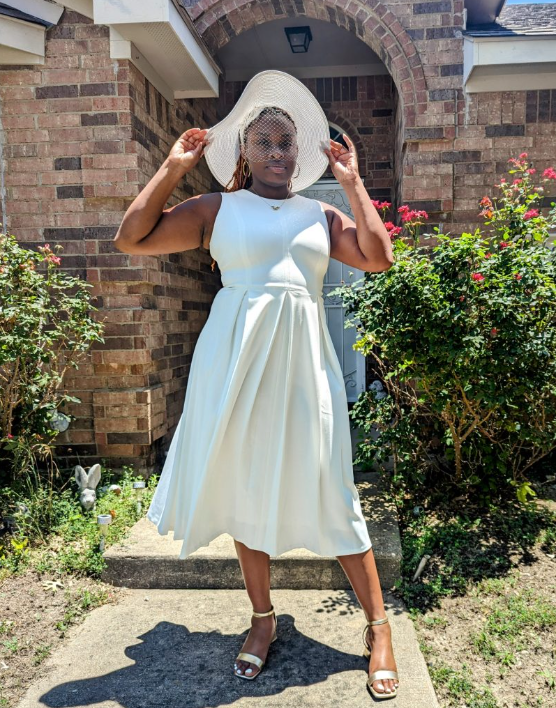 NeNe stands in the walkway of a familial home dressed in a white dress, large white hat, short white veil, and gold heels. Her expression is somber as she prepares to attend her father's funeral.
