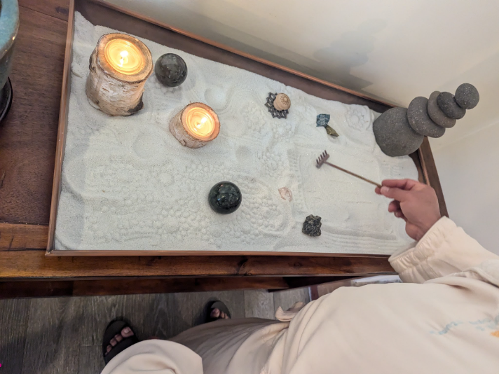 A person in white clothing uses a small wooden rake to create patterns in a tabletop Zen sand garden filled with white sand, stones, crystals, and two lit candles. The calming arrangement sits on a wooden table and evokes meditation, mindfulness, and intentional rest.