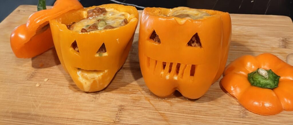 There are 2 jack o lantern stuffed bell peppers with different expressions. One has cheese flowing out of its mouth.