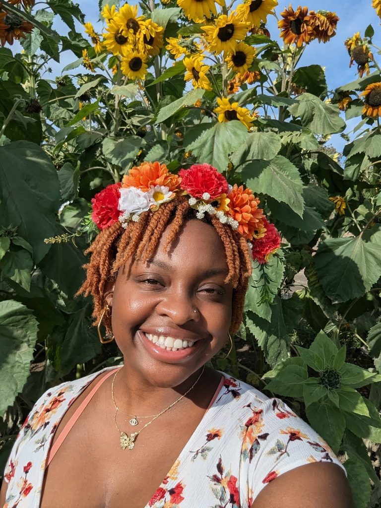 NeNe is smiling in the front of large clusters of sunflowers at a local pumpkin patch. She is dressed in autumn florals and has a flower headband on.